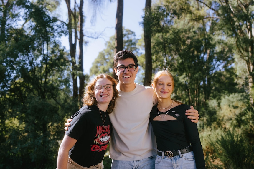 A young man has his arms around two young women as they stand in front of some trees