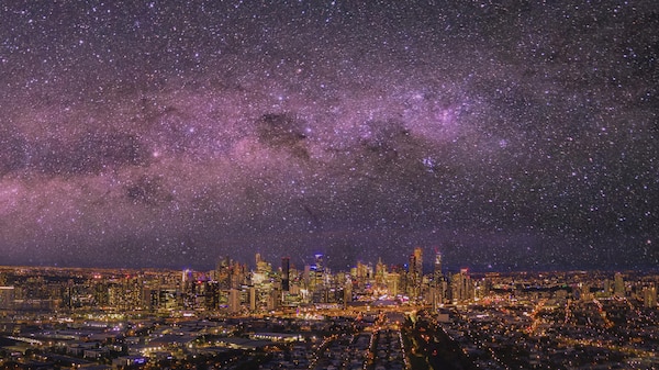 Above a city landscape the Milky Way appears as a pinkish swathe of stars across the night sky.