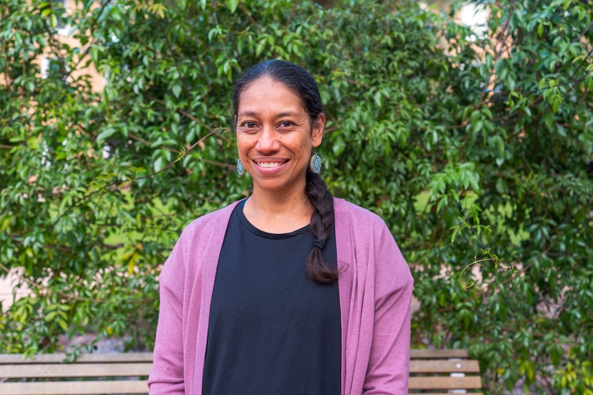 A woman smiling with a long black plait wearing a pink cardigan in front of green shrubbery.