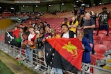 A group of rugby league fans stand by the railing in front of a half-empty stand, holding PNG flags. 