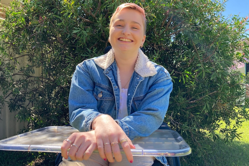 A young woman looks to the camera and smiles with her hands crossed in front of her on her wheelchair. She wears a jean jacket.