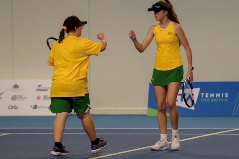 Two woman wearing Australian gold shirts and green skirts give each other a fist pump on the tennis court.