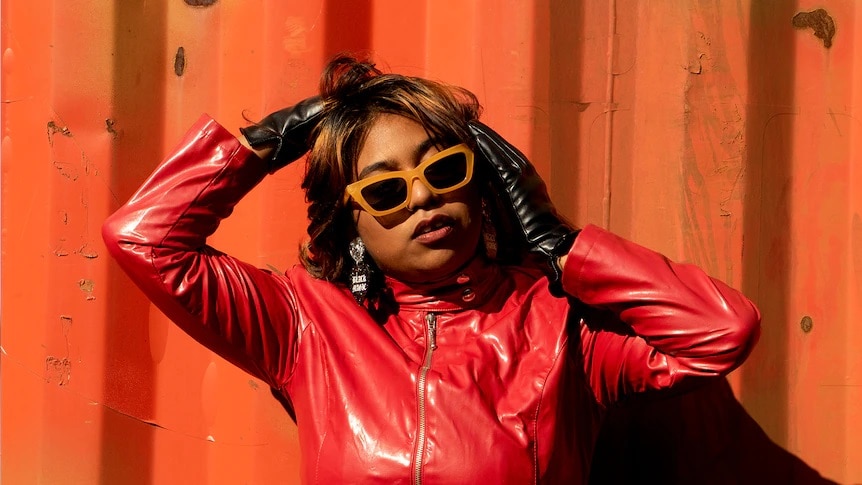 A woman wearing sunnies and a red jacket against an orange wall outside