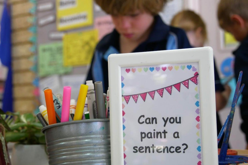 A small white sign on a table in a classroom reads "can you paint a sentence?", in front of a young boy standing up.