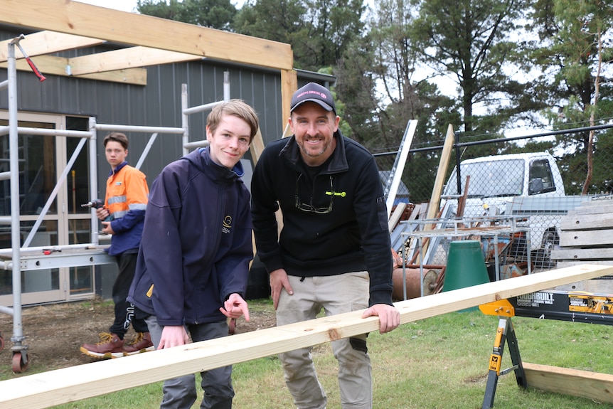 Harper Ross and Dave Colcott smile for the camera while leaning over a plank of wood behind a renovation project.