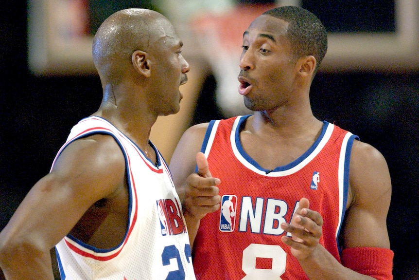 Michael Jordan in a white basketball jersey talks to Kobe Bryant in a red jersey