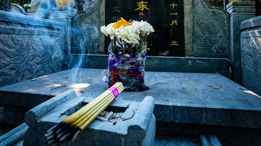 Yellow and white flowers and a sticks of burning incense next to a black headstone with Chinese characters written on it