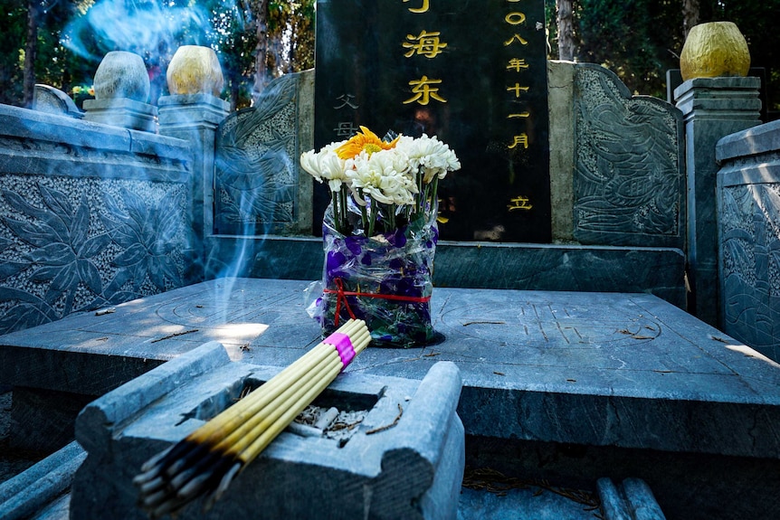 Yellow and white flowers and a sticks of burning incense next to a black headstone with Chinese characters written on it