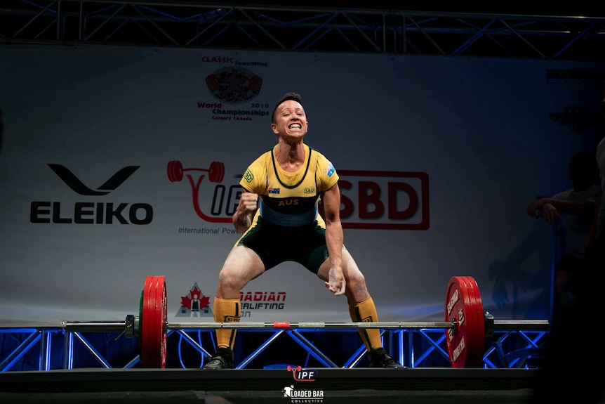 A woman powerlifter celebrates with weights in front of her, she is wearing Australian representative clothing.