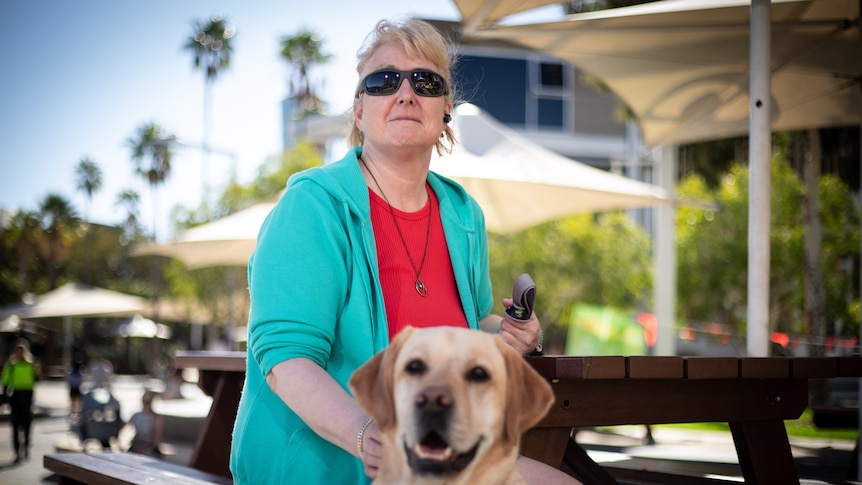 Woman with short blond hair sitting outdoors with her guide dog in front of her