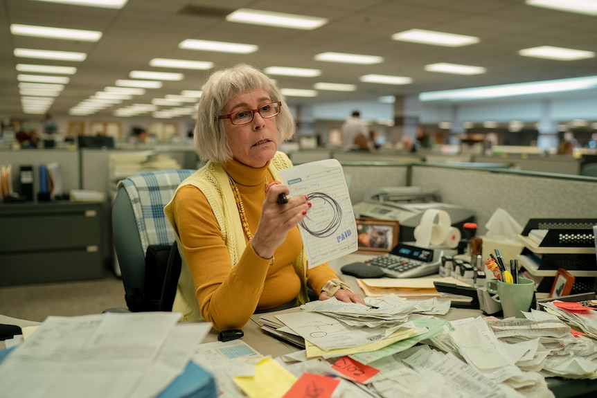 White woman with scraggly short white hair, red glasses, mustard skivvy and weary expression holds up document in dreary office.