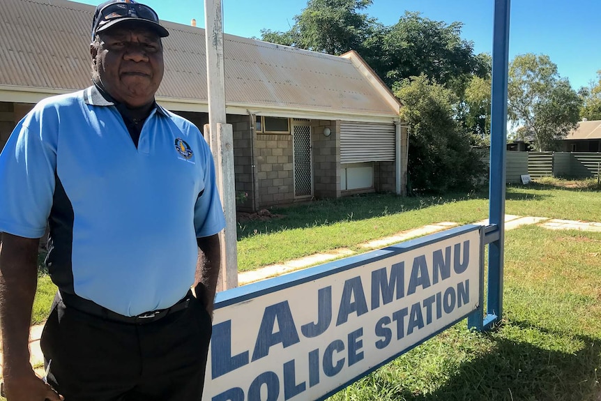 An Indigenous man wearing a blue police liaison officer's polo shirt stands at the front of a single-level block building.