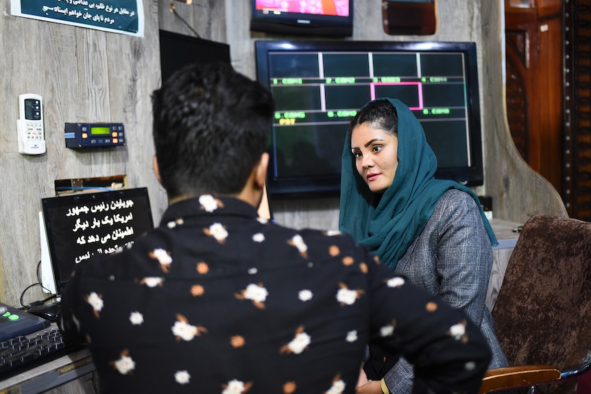 A young woman sits in a studio type room, surrounded by TV screens