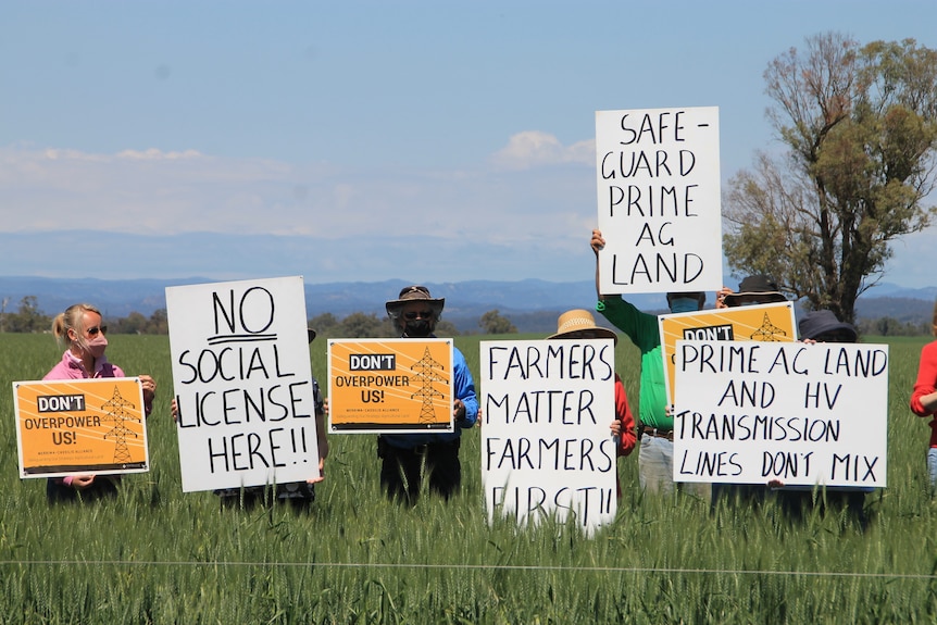 A group of farmers stand in a paddock holding protest signs like "Farmers matter, farmers first".