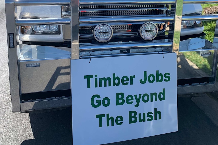 A freight truck is parked on the road on a sunny day with a sign attached to the front that says: Timber Jobs Go Beyond The Bush