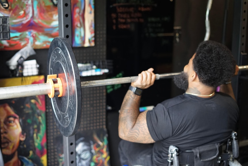 Man in wheelchair holding heavy barbells wearing black tee