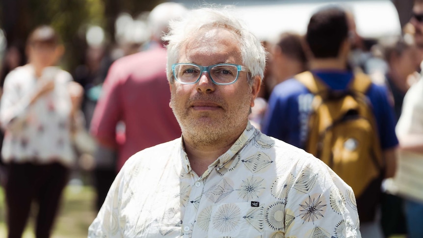 Gerd Shroder-Turk, wearing short-sleeve collared shirt and blue rimmed glasses, stands outside with crowd visible in background.