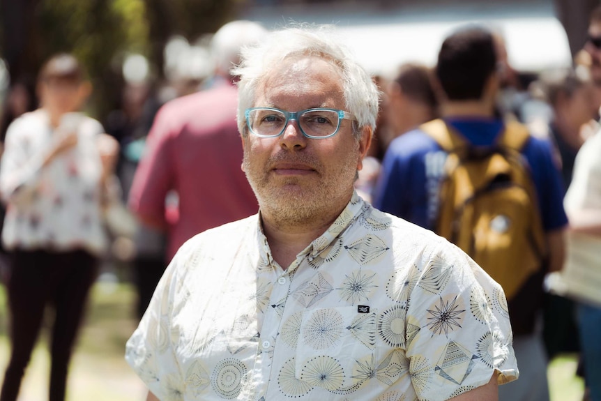 Gerd Shroder-Turk, wearing short-sleeve collared shirt and blue rimmed glasses, stands ouside with crowd visible in backgrounnd.