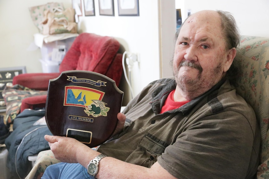 Man sitting in armchair holding up shield trophy featuring map of SA and fish 