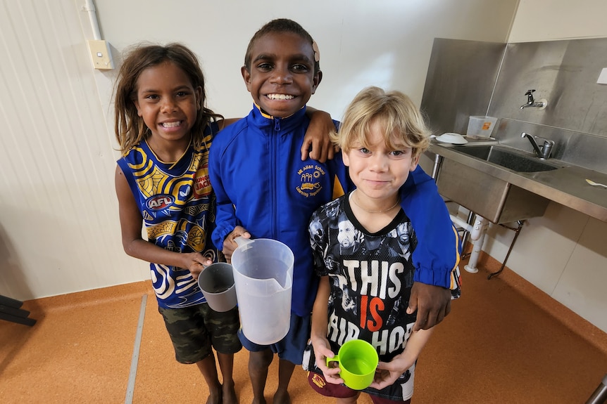 three primary school children standing with arms around each other, smiling. Each is holding a cup or container for water