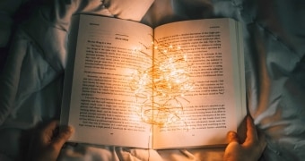 Open book on a bedspread, with hands. On the book a tangle of lights. Book is illuminated