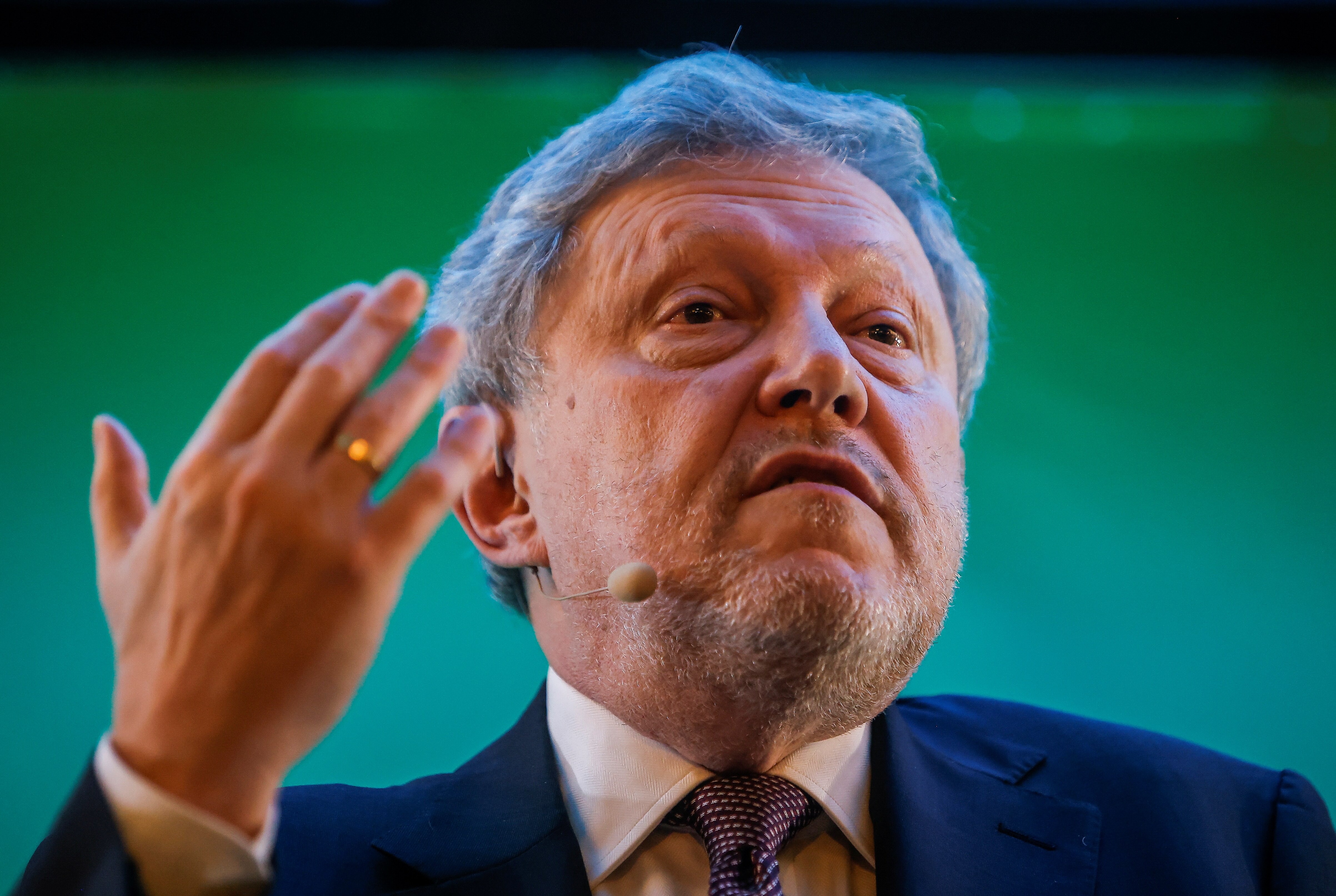 An older man in a suit gestures during a speech with a small mic attached to his face 