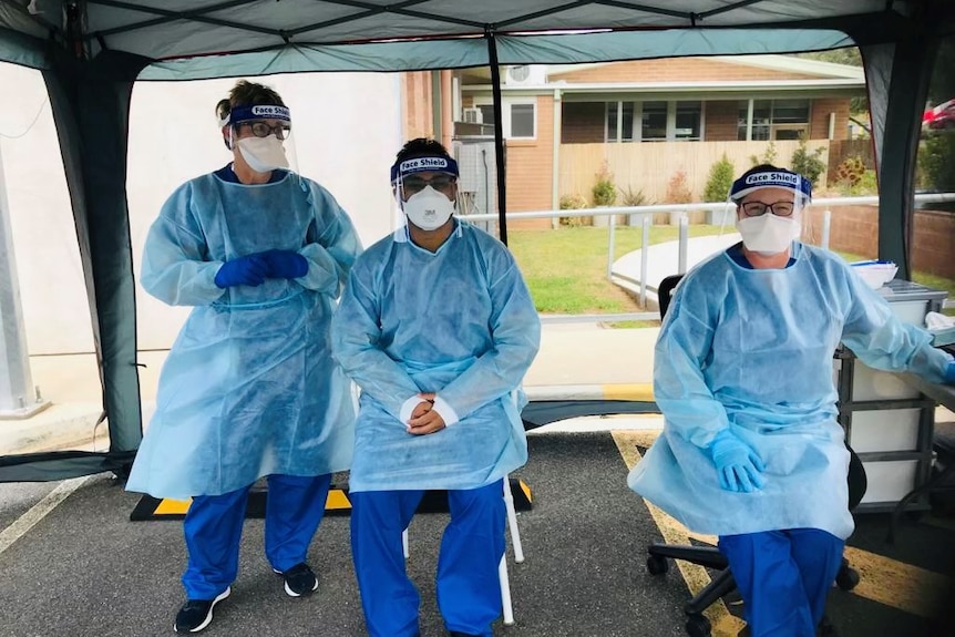 Three people in blue gowns, face shield and masks sitting under a tent.