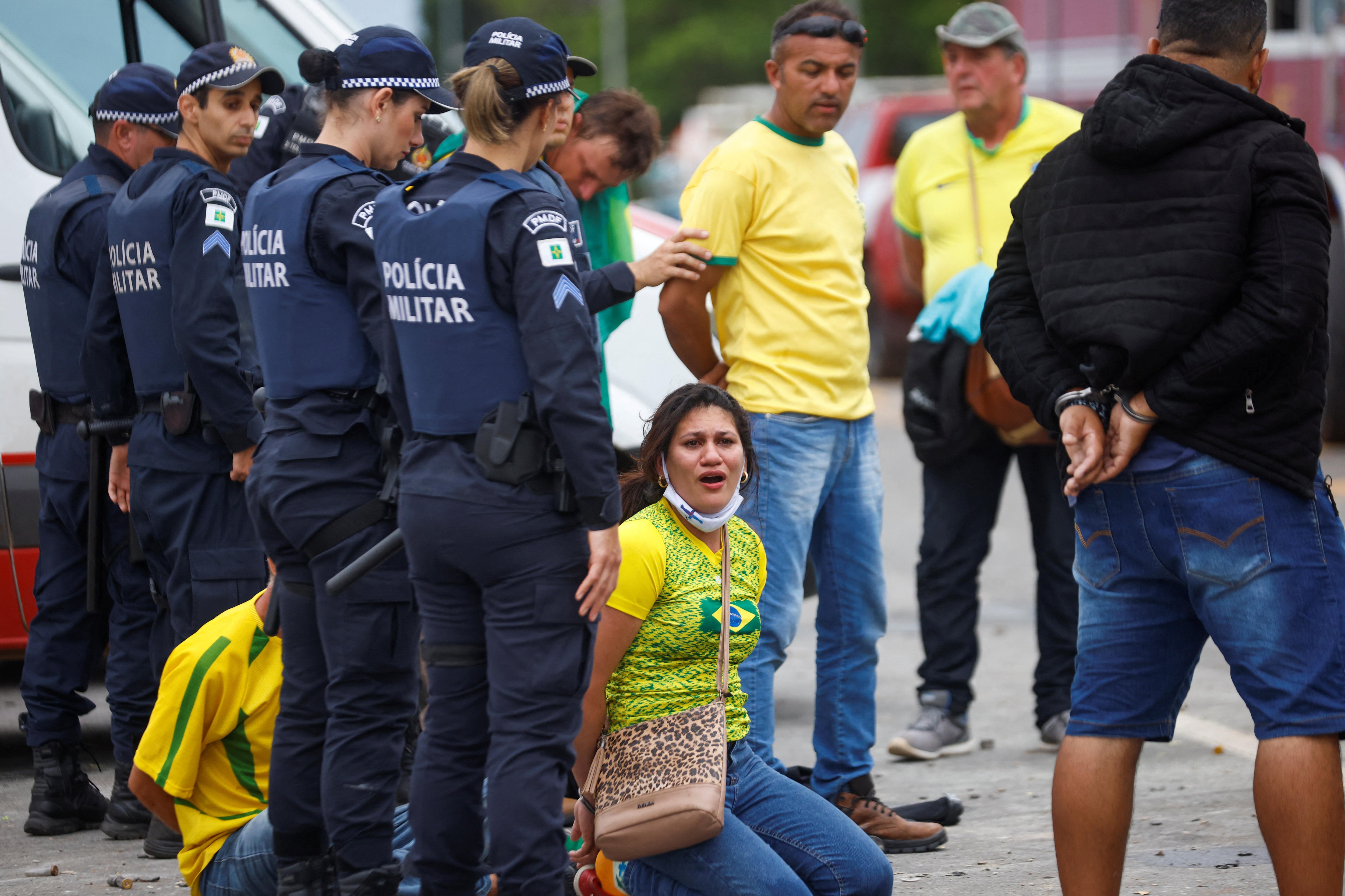 A woman wearing a yellow tshirt sits on the ground with her hands tied behind her back surrounded by police.