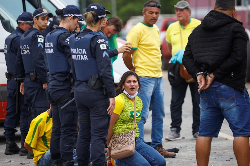 A woman wearing a yellow tshirt sits on the ground with her hands tied behind her back surrounded by police.