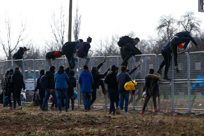 A line of migrants climb a fence as others wait behind them.