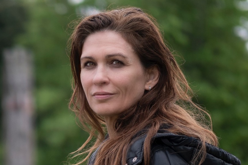 Elissa Down, with long auburn hair and black puffer jacket, stands turning head sideways towards camera, smiling slightly.