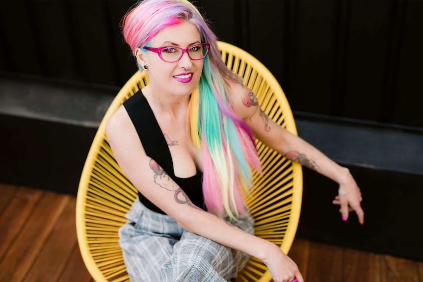 Woman with pink and teal hair, wearing glasses, sitting in yellow chair. 