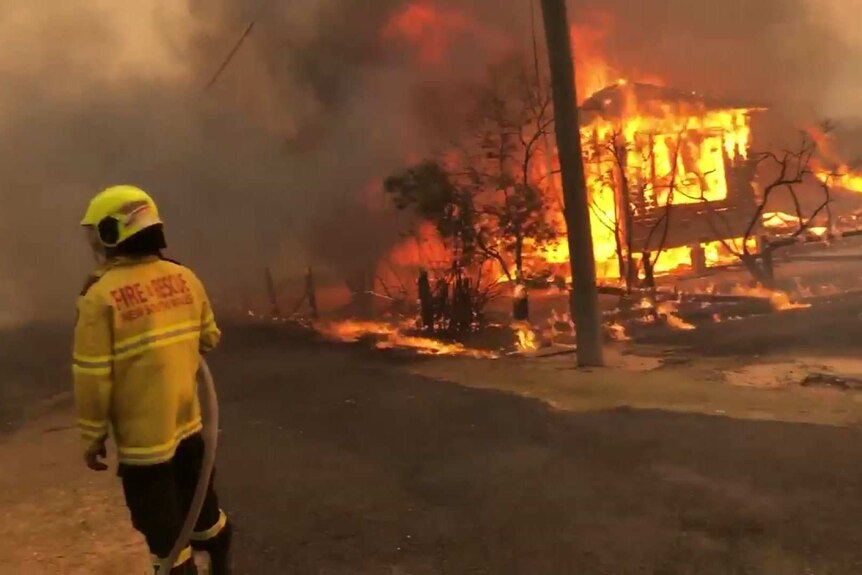 A firefighter watches as a building burns in front of a smoky background.