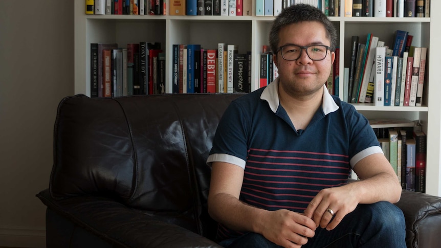 A 31-year-old Asian man sits on a brown leather armchair in front of a book case