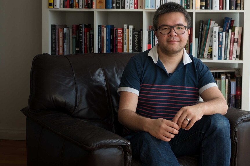 A 31-year-old Asian man sits on a brown leather armchair in front of a book case
