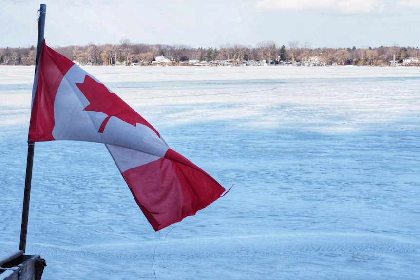 Frozen snowy lake in Canada with Canadian flag in the foreground and houses and trees in the background.