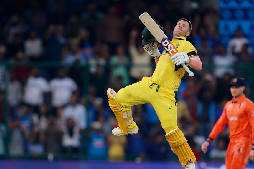 A young man in a yellow and green cricket uniform leaps in the air with his helmet off in triumph on a cricket field. 
