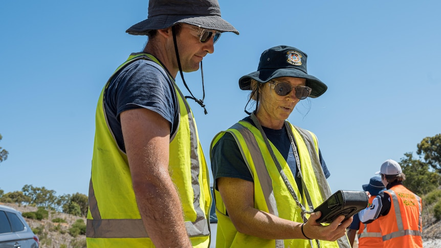 Two people in high vis clothing look at a small black box with a screen on a sunny day.