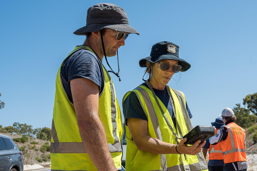 Two people in high vis clothing look at a small black box with a screen on a sunny day.
