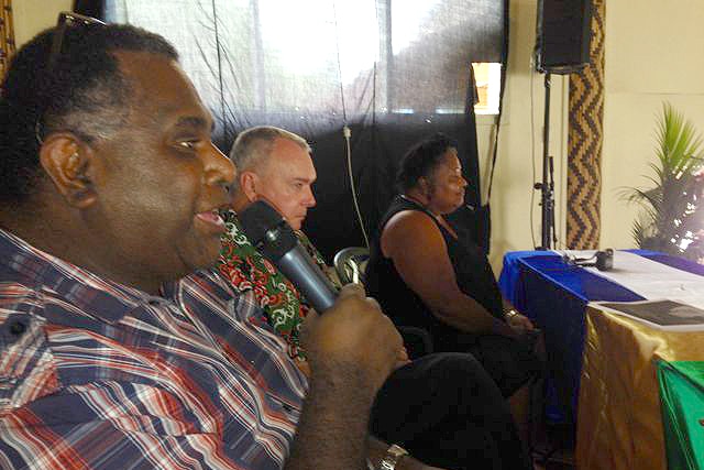 Man of South Sea Islander heritage holds mic sitting at table by another man and woman.