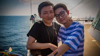 Two men stand smiling with their arms around each other on the deck of a cruise ship.