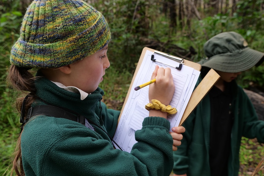 A primary school girl wearing a green top and a beanie writes notes on a clipboard in a bushland area.