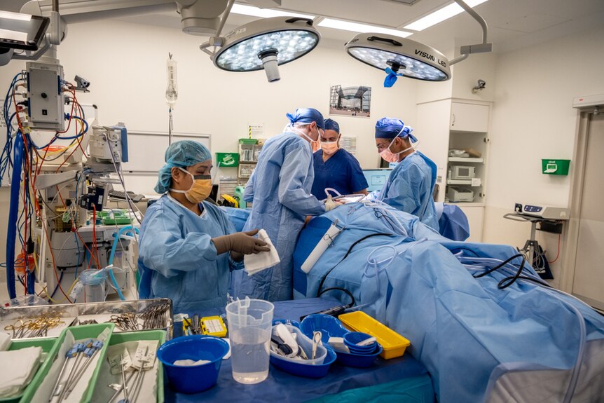 Surgeons lean over an obscured woman in an operating theatre as they perform surgery on her breasts.