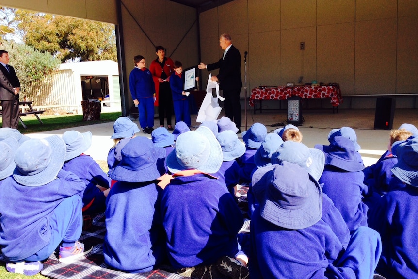 A sea of blue uniforms watch on as two students and a principal hold a certificate in an outdoor award ceremony