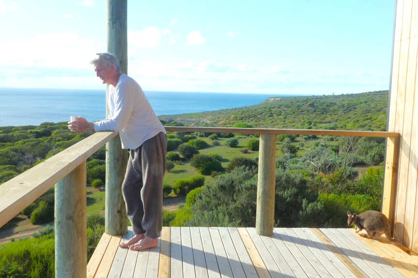 Man leaning on balcony railing with coffee cup, looking out to sea, small wallaby marsupial behind him, coastal background