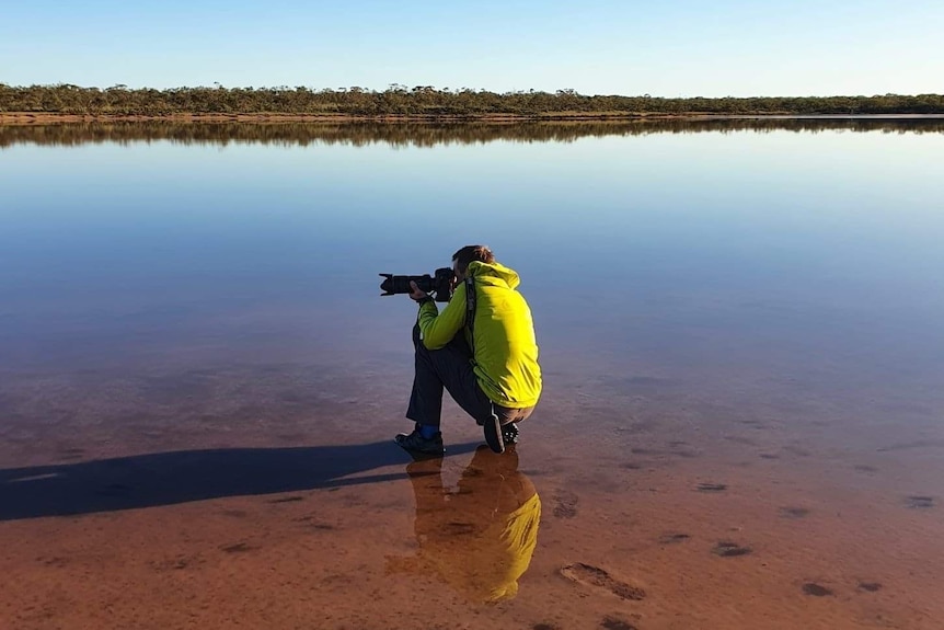 A man in a yellow parker kneels in shallow water to take a landscape photo.
