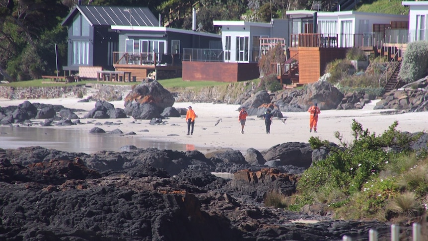 Four people walk along a beach in front of shacks.