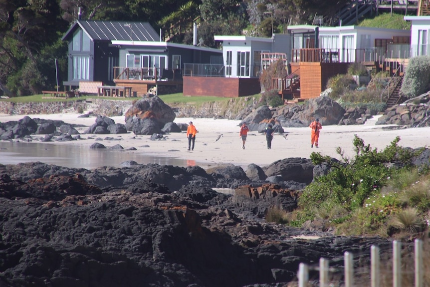 Four people walk along a beach in front of shacks.