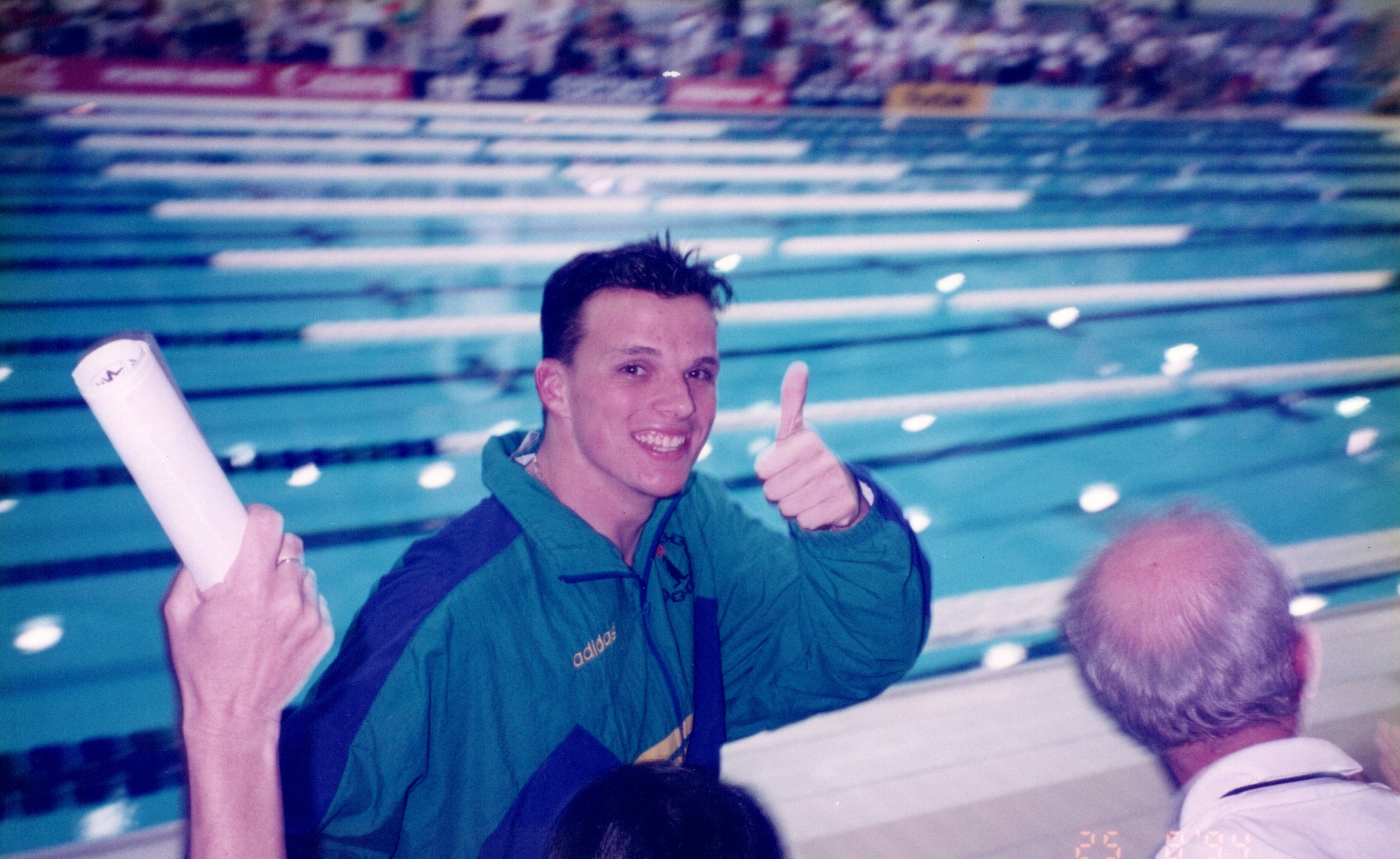 A young Scott Miller wearing an Australian green and gold tracksuit giving the thumbs up in front of a swimming pool.