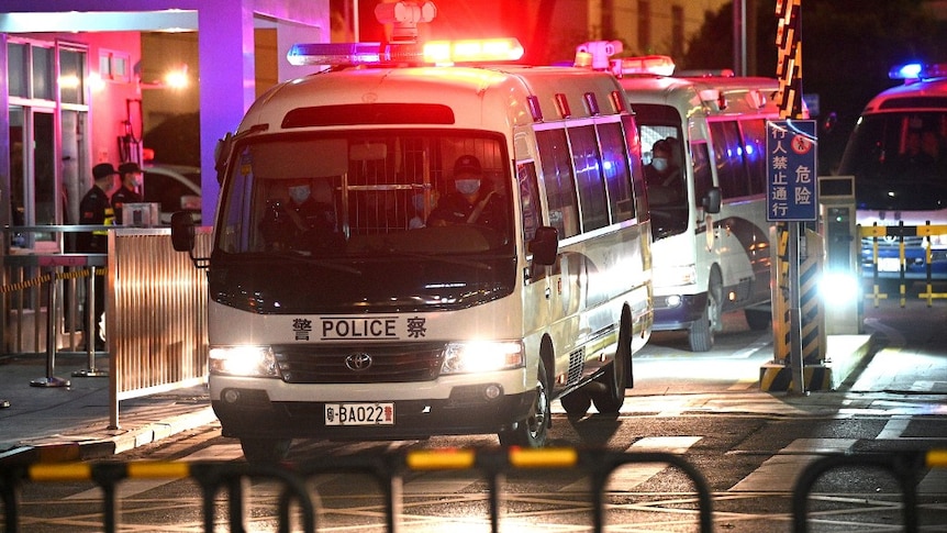 At night, you view a convoy of Chinese police vans as with the street bathed in blue, red, and white light.